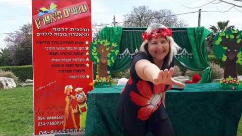 המדריכה על במת הגן, לצד תפאורת עצים ופרחים ושלט התוכנית